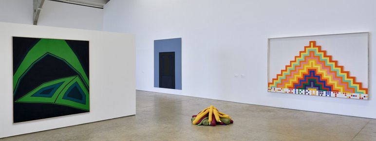 An art gallery interior featuring three artworks. On the left, a large dark green abstract painting; in the centre, a colourful geometric sculpture resembling a campfire; on the right, a vibrant triangular painting with stripes. The space is bright and minimalistic.