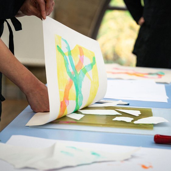 A person in a grey sweater is lifting a sheet of paper from a table covered with various artworks and tools. The background features colourful abstract designs, while the table holds additional sheets and a roller.