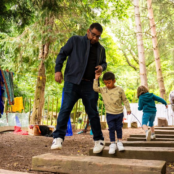 A man helps a young child navigate wooden steps in a forested area. The child is wearing a light-coloured top and navy trousers. In the background, other children and adults are engaged in activities among trees and hanging clothes.