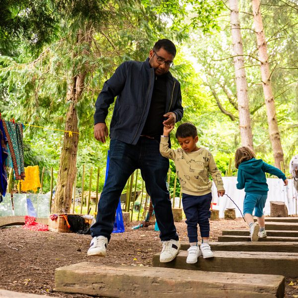 A man helps a young child navigate wooden steps in a forested area. The child is wearing a light-coloured top and navy trousers. In the background, other children and adults are engaged in activities among trees and hanging clothes.