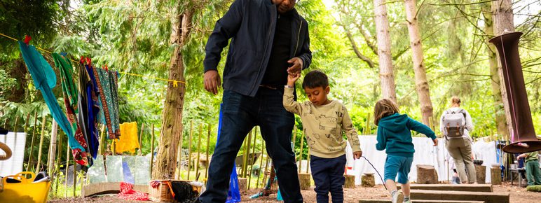 A man helps a young child navigate wooden steps in a forested area. The child is wearing a light-coloured top and navy trousers. In the background, other children and adults are engaged in activities among trees and hanging clothes.