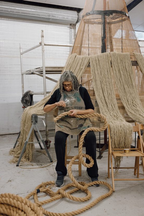 A person with long, grey hair sits on a chair in a workshop, carefully working with thick ropes. Surrounding them are large, woven structures and tools, creating a creative and industrious atmosphere. Natural light filters through the space.