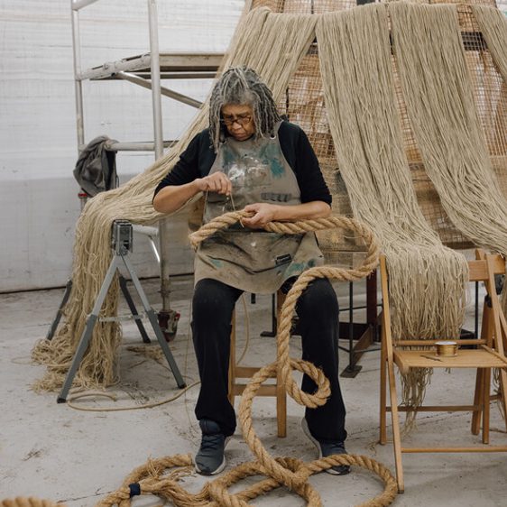 A person with long, grey hair sits on a chair in a workshop, carefully working with thick ropes. Surrounding them are large, woven structures and tools, creating a creative and industrious atmosphere. Natural light filters through the space.