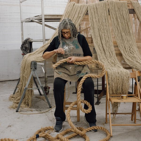 A person with long, grey hair sits on a chair in a workshop, carefully working with thick ropes. Surrounding them are large, woven structures and tools, creating a creative and industrious atmosphere. Natural light filters through the space.