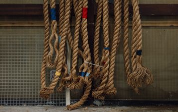 A collection of thick, natural fibre ropes hanging against a textured background. The ropes are tied with colourful bands of red, blue, and yellow, showcasing various lengths and frayed ends, creating a rustic and industrial aesthetic.
