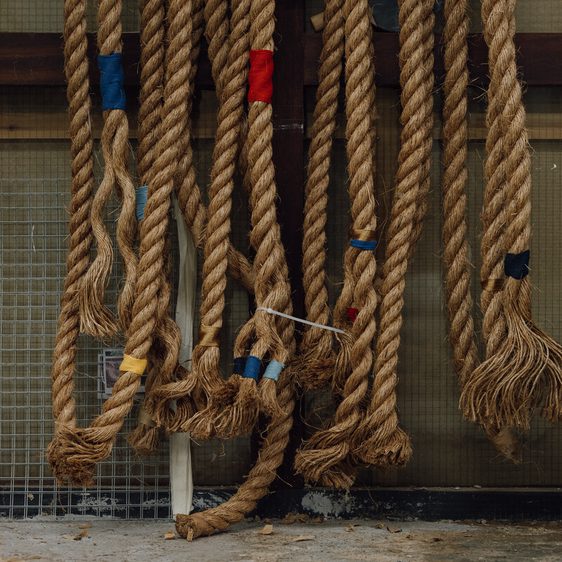 A collection of thick, natural fibre ropes hanging against a textured background. The ropes are tied with colourful bands of red, blue, and yellow, showcasing various lengths and frayed ends, creating a rustic and industrial aesthetic.