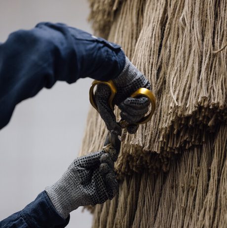 A close-up of hands wearing grey gloves, holding golden scissors, trimming a large bundle of natural fibre strands. The background features a textured wall of tightly packed, hanging fibres, creating a warm, earthy atmosphere.