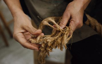 A close-up of two hands skillfully working with natural fibres, twisting and shaping them. The background is softly blurred, focusing on the intricate details of the fibres and the hands, suggesting a traditional craft or weaving process.