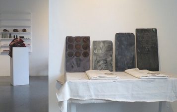 An art installation featuring a large black-and-white mural of a tree, a wooden bench, and various sculptures. The space includes a white pedestal displaying a red fabric piece and photographs arranged on a nearby shelf.