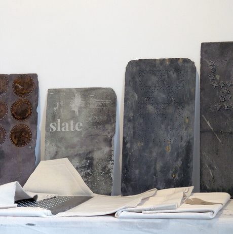 Four pieces of slate, with stitched experimental sections lean against a white wall. other pieces of slate, wrapped in white fabric, are displayed on a shelf in front of them.