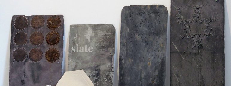 Four pieces of slate, with stitched experimental sections lean against a white wall. other pieces of slate, wrapped in white fabric, are displayed on a shelf in front of them.