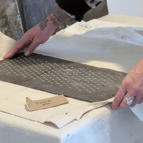 An art installation featuring a large black-and-white mural of a tree, a wooden bench, and various sculptures. The space includes a white pedestal displaying a red fabric piece and photographs arranged on a nearby shelf.