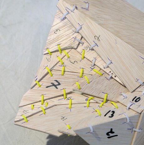 An art installation featuring a large black-and-white mural of a tree, a wooden bench, and various sculptures. The space includes a white pedestal displaying a red fabric piece and photographs arranged on a nearby shelf.