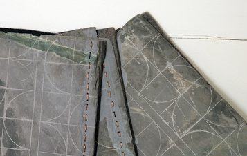 An art installation featuring a large black-and-white mural of a tree, a wooden bench, and various sculptures. The space includes a white pedestal displaying a red fabric piece and photographs arranged on a nearby shelf.