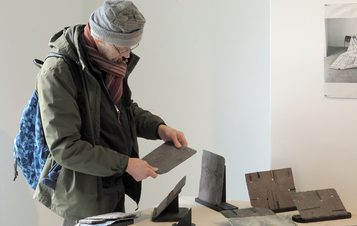 An art installation featuring a large black-and-white mural of a tree, a wooden bench, and various sculptures. The space includes a white pedestal displaying a red fabric piece and photographs arranged on a nearby shelf.