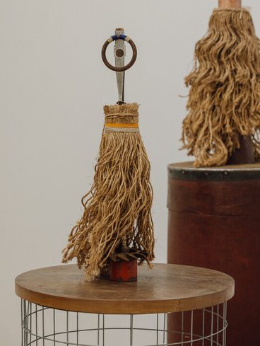 A decorative object resembling a mop, made of natural fibres, stands on a wooden table. It features a metal handle with a circular ring at the top. In the background, a similar object is displayed on a cylindrical red base.