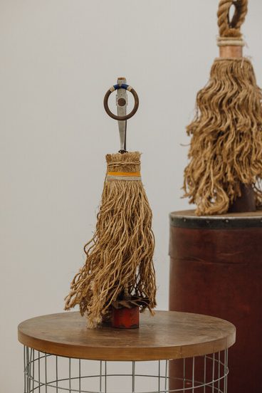 A decorative object resembling a mop, made of natural fibres, stands on a wooden table. It features a metal handle with a circular ring at the top. In the background, a similar object is displayed on a cylindrical red base.