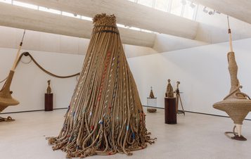 An art installation featuring large, intricately woven rope sculptures in a bright, modern gallery. The central piece resembles a conical shape, surrounded by smaller sculptures on pedestals, with natural light streaming through the skylights above.