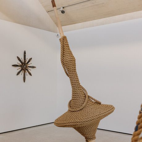 A large, sculptural installation made of thick, coiled rope hangs from the ceiling, resembling a spinning top. The minimalist gallery space features a white wall with a star-shaped artwork in the background, enhancing the contemporary aesthetic.