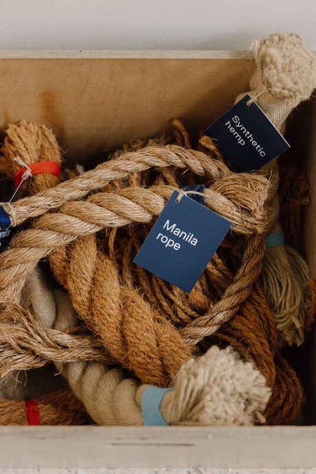 A wooden box filled with various types of rope, including manila rope and synthetic hemp. Each rope is neatly coiled and labelled with blue tags indicating their type. The textures of the ropes are visible, showcasing their natural fibres.
