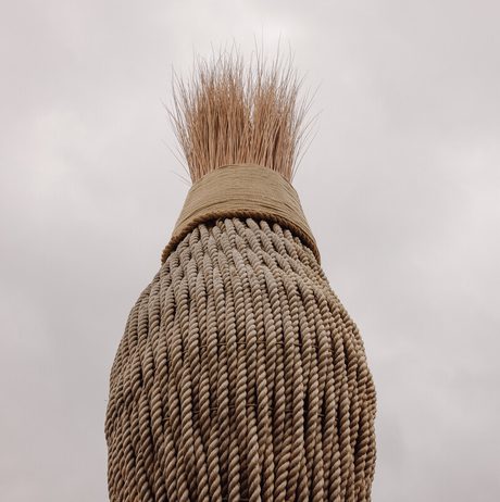 A large, textured broom with a thick, woven handle and a bushy bristle head stands against a cloudy sky. The focus is on the broom's intricate details and natural materials, creating a striking silhouette.