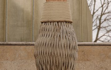 A large, artistic broom made of natural fibres stands against a textured wall. The broom's bristles are bushy and light, contrasting with the earthy tones of the wall and the muted background of trees and a modern building.