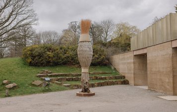 A large, artistic broom sculpture stands in a gravel area, surrounded by greenery and stone features. The broom has a woven base and a bushy bristle top, contrasting with the modern building in the background under a cloudy sky.