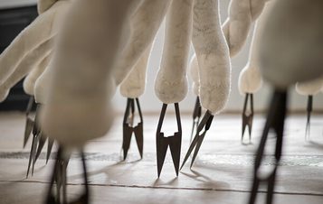 A close-up view of an abstract sculpture featuring elongated, soft, white forms resembling tentacles. Each tentacle ends in sharp, metallic points, creating a striking contrast between softness and danger, set against a minimalist background.