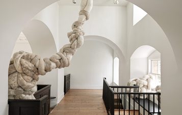 An interior space featuring a large, soft sculpture made of intertwined, plush materials. The sculpture extends from the upper level, curving through an archway, with white walls and wooden flooring creating a minimalist backdrop.