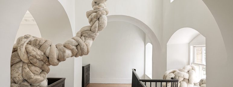 An interior space featuring a large, soft sculpture made of intertwined, plush materials. The sculpture extends from the upper level, curving through an archway, with white walls and wooden flooring creating a minimalist backdrop.