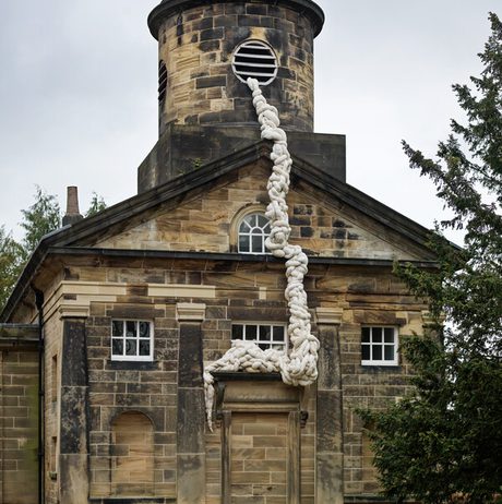 A historic stone building with a round tower features a large, white, rope-like installation cascading down its side. The structure has multiple windows and a door, surrounded by greenery and a cloudy sky above.