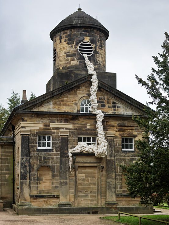 A historic stone building with a round tower features a large, white, rope-like installation cascading down its side. The structure has multiple windows and a door, surrounded by greenery and a cloudy sky above.
