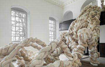 A large, abstract sculpture made of intertwined, soft, rope-like forms occupies a bright, minimalist room. The sculpture's organic shapes contrast with the clean lines of the architecture, while natural light filters through large windows.