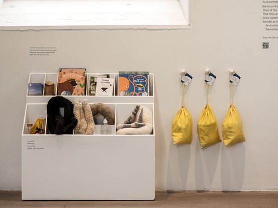 A white display unit holds various items, including books and plush toys. Beside it, three yellow bags hang from hooks on the wall. The setting is bright and minimalistic, with a large window providing natural light.