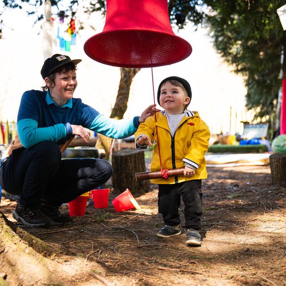 A smiling adult in a blue top and cap interacts with a young child wearing a yellow jacket and black hat. The child holds a wooden mallet, standing under a large red bell hanging from a tree, surrounded by a playful outdoor setting.