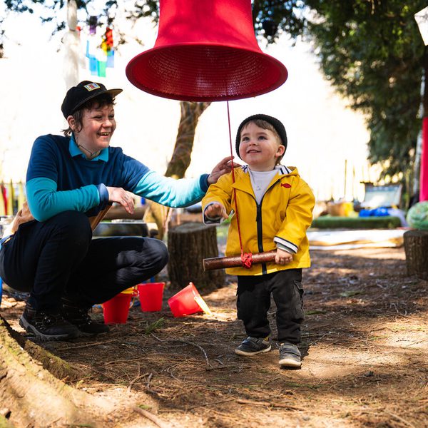 A smiling adult in a blue top and cap interacts with a young child wearing a yellow jacket and black hat. The child holds a wooden mallet, standing under a large red bell hanging from a tree, surrounded by a playful outdoor setting.