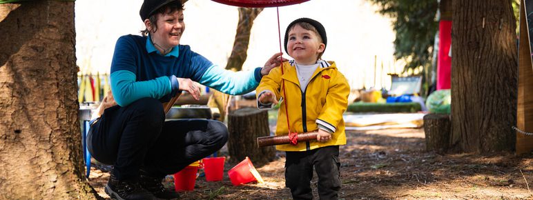 A smiling adult in a blue top and cap interacts with a young child wearing a yellow jacket and black hat. The child holds a wooden mallet, standing under a large red bell hanging from a tree, surrounded by a playful outdoor setting.