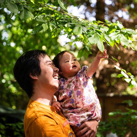 A man in an orange shirt holds a smiling child wearing a floral outfit. They are outdoors, surrounded by lush green leaves. The child is reaching up towards the branches, both appearing joyful and engaged with nature.