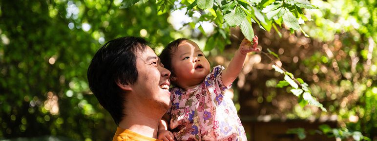 A man in an orange shirt holds a smiling child wearing a floral outfit. They are outdoors, surrounded by lush green leaves. The child is reaching up towards the branches, both appearing joyful and engaged with nature.