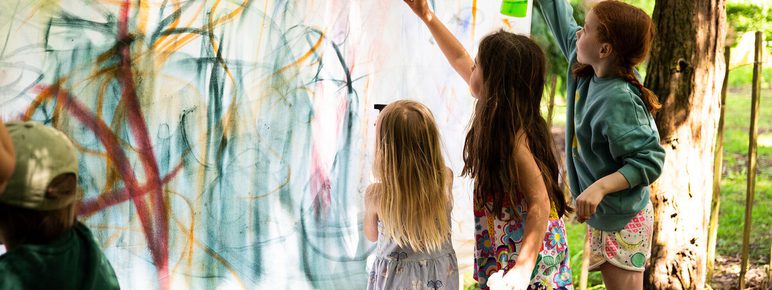 Children painting on a large canvas outdoors, using spray bottles and brushes. The canvas is filled with colourful abstract lines and shapes. Sunlight filters through the trees, creating a lively and creative atmosphere.