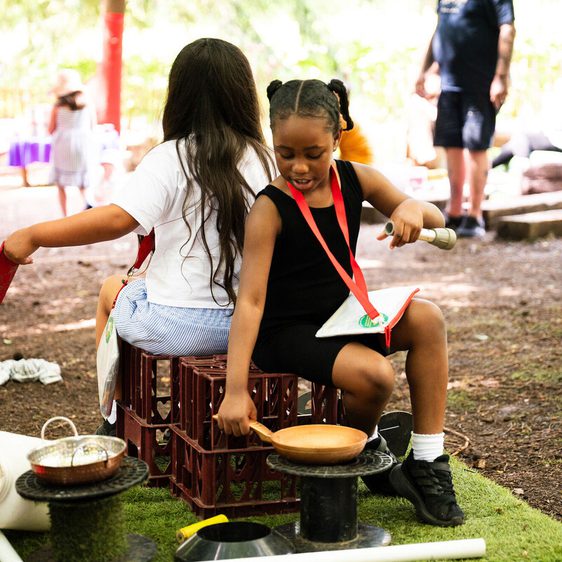 Two children sit back-to-back on crates outdoors, playing with kitchen utensils. One holds a pan and spoon, while the other has a red board. Various objects are scattered around. Adults and other children are visible in the background.