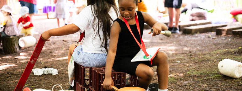 Two children sit back-to-back on crates outdoors, playing with kitchen utensils. One holds a pan and spoon, while the other has a red board. Various objects are scattered around. Adults and other children are visible in the background.