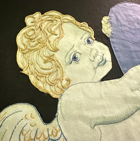 A close-up of an embroidered cherub with golden curls and blue eyes on a dark background. The cherub has white wings with gold accents and is partially holding a light purple object. The stitching is detailed and textured.