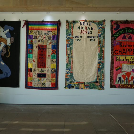 A display of four colourful quilt panels commemorating individuals affected by AIDS. Each panel features names, dates, and decorative elements, such as patterns and symbols, against a white wall in a well-lit gallery space.