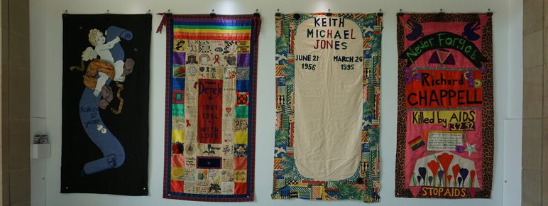 A display of four colourful quilt panels commemorating individuals affected by AIDS. Each panel features names, dates, and decorative elements, such as patterns and symbols, against a white wall in a well-lit gallery space.