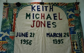 A colourful quilt panel with the name "Keith Michael Jones" in bold letters. Dates "June 21, 1956" and "March 26, 1995" are displayed below. The background features vibrant, abstract patterns in various colours.