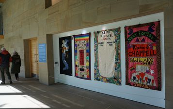 A hallway display features colourful quilt panels commemorating individuals affected by AIDS. Each panel is uniquely designed with names, dates, and vibrant patterns. Two people stand nearby, viewing the exhibit.