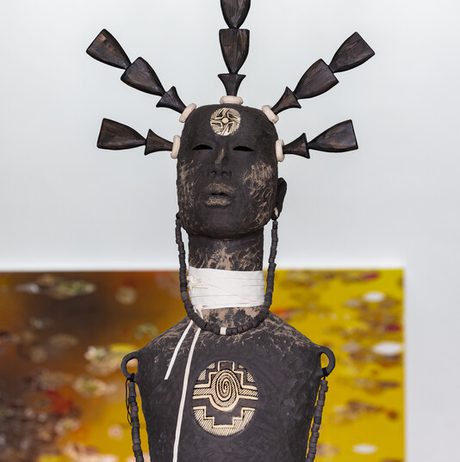 A sculpture of a figure with a textured, dark surface, adorned with geometric patterns. The head features multiple upward-pointing elements. The background shows blurred, colourful artwork on a white wall.