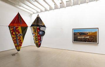 Two large kites with bold text hang in a white gallery. A framed photograph on the right shows a desert landscape with "INDIAN LAND" written on it. The ceiling features exposed beams and the floor is light-coloured.