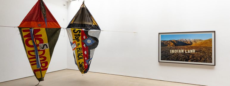 Two large kites with bold text hang in a white gallery. A framed photograph on the right shows a desert landscape with "INDIAN LAND" written on it. The ceiling features exposed beams and the floor is light-coloured.